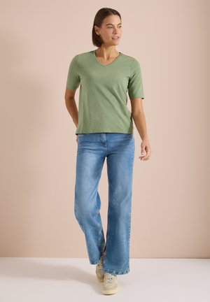 Femme portant un t-shirt vert à manches courtes et col en V, un jean bleu à jambes larges et des baskets blanches, debout sur un fond beige.