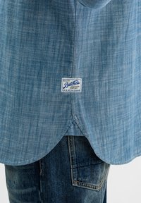 Lichtblauw overhemd met korte mouwen en een getailleerde zoom, gemaakt van gestructureerde stof. Heeft een wit label met blauwe tekst op de achterkant.