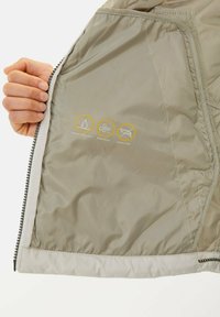 Main tenant une veste beige clair ouverte montrant l'intérieur avec des icônes des caractéristiques déperlantes, coupe-vent et sans duvet.