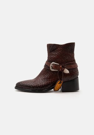 Bottine en cuir tressé marron foncé avec un talon bas et épais, une bride à boucle argentée et une étiquette de marque orange attachée sur fond uni.