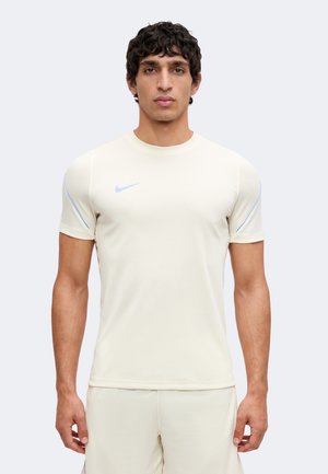 Muž stojaci čelom vpred, oblečený v priliehavé svetlobéžové športové tričko Nike s krátkymi rukávmi a zodpovedajúce šortky.