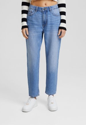 Frau trägt hellblaue Jeans mit geradem Bein, weiße Sneakers und einen schwarz-weiß gestreiften, langärmeligen, kurzen Pullover.