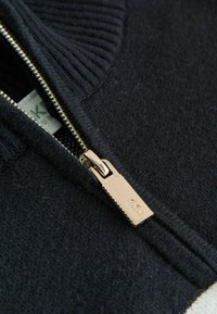 Suéter azul marino con cuello acanalado, presenta una cremallera dorada y un tirador. Textura suave, mostrando un primer plano del área de la cremallera.