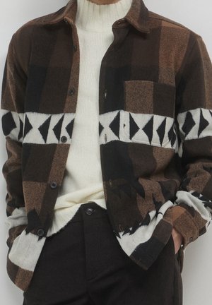 Personne portant une chemise à carreaux marron et noir avec un motif géométrique blanc, sur un pull à col roulé crème et un pantalon noir.