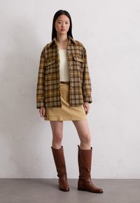 Bruin geruite oversized shirt, beige tanktop, kaki minirok en lange bruine leren laarzen. Heeft twee borstzakken en een ceintuur in de taille.