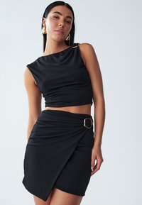 Černý zkrácený top s jedním ramínkem, střižený design. Černá zavinovací sukně s detailem zlaté čtvercové spony. Hladká textura, přiléhavý silueta.