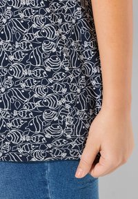 Tessuto navy con un motivo di pesci bianchi e forme geometriche. Primo piano di una mano che tiene il bordo del capo, rivelando la texture e il design.