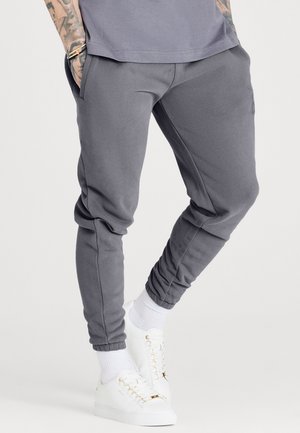 Mand iført grå slim-fit joggingbukser, hvide sokker og hvide sneakers, stående mod en ensfarvet baggrund.