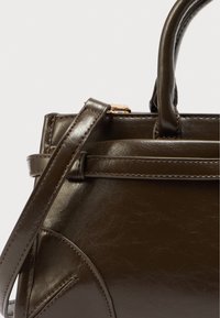 Σκούρα καφέ δερμάτινη τσάντα χειρός με δομημένο σχεδιασμό, διπλές λαβές και αποσπώμενο λουρί. Διαθέτει διακριτικές ραφές και χρυσές μεταλλικές λεπτομέρειες.