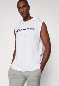 Balta be rankovių sporty hoodie su raišteliu ir tamsiai mėlynu "Champion" logotipu. Dėvima su pilkomis sportinėmis kelnėmis, turinčiomis mažą logotipą ant šlaunies.