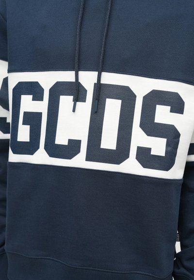 Sötétkék és fehér kapucnis pulcsi, merész, sötétkék "GCDS" felirattal a mellrészen, és azonos színű zsinórok lógnak lefelé.