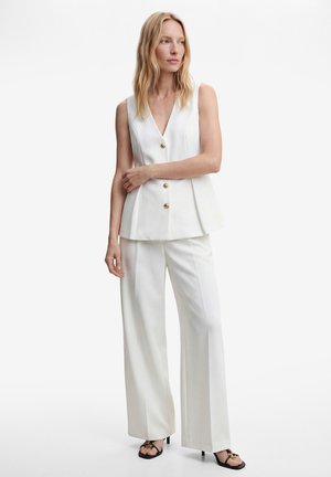 Frau trägt weiße ärmellose, geknöpfte Weste und weite Hosen mit schwarzen Sandalen mit Absätzen, stehend vor einfarbigem Hintergrund.