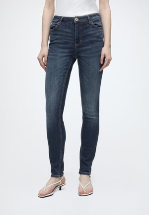 Persoon die donkerblauwe skinny jeans en witte sandaaltjes met lage hak en bandjes draagt, staand tegen een effen witte achtergrond.