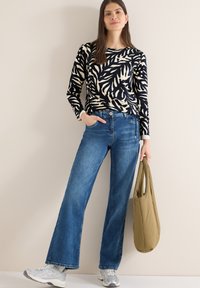 Frau steht vor einer beigen Wand, trägt ein langärmeliges Oberteil mit schwarz-weißem Blättermuster, blaue Jeans mit weitem Bein, weiße Turnschuhe und hält eine olivgrüne Tragetasche.