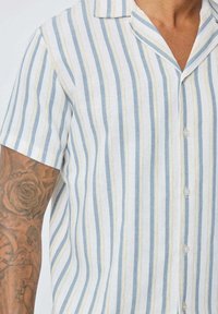 Chemise en lin à manches courtes avec des rayures verticales bleu clair et blanches, dotée d'une texture douce, d'un col et d'une fermeture à boutons.