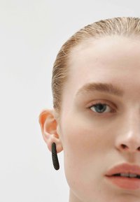 Pendiente de aro texturizado negro que se lleva en la oreja izquierda. El pendiente presenta una forma cilíndrica con un acabado mate y un detalle sutil.