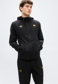 Μαύρο ελαφρύ μπουφάν με κουκούλα, με χρυσά λογότυπα στο στήθος και στα μανίκια, σχεδιασμένο με ομαλή υφή και φερμουάρ κλεισίματος.