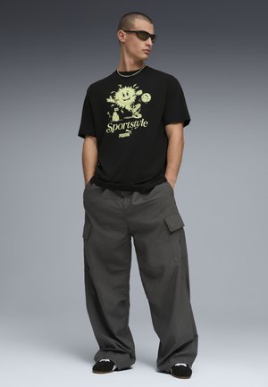 Hombre con camiseta negra "Sportstyle Puma" con sol de dibujos animados y balón de fútbol, pantalones cargo grises, gafas de sol negras y zapatos negros, con las manos en los bolsillos.