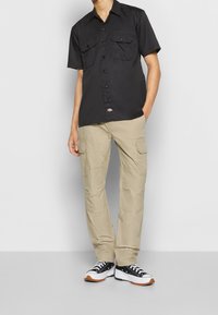 Kortärmad svart button-up skjorta med två bröstfickor, i kombination med beige cargobyxor som har flera fickor och svarta sneakers.