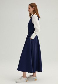 Robe bleu marine avec un corsage ajusté et une jupe évasée. Chemise blanche à manches longues en dessous, complétée par une texture douce et des poches.