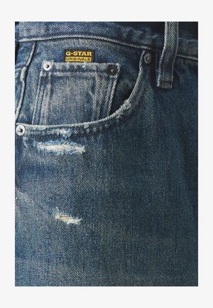 A G-Star farmernadrág kék színű, elmosódott mintázatú, koptatott részletekkel rendelkezik, és a zsebében látható a márka címkéje.