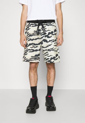 Mężczyzna w czarno-białych szortach o abstrakcyjnym wzorze z czarnym sznurkiem, czarnych skarpetkach, czarnych tenisówkach i białej koszulce, stojący.