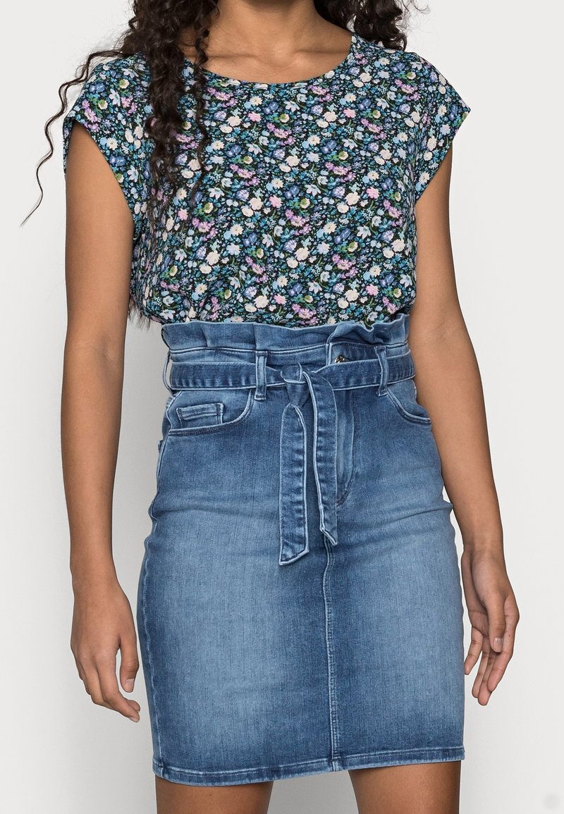 Personne portant un haut à manches courtes à motif floral rentré dans une jupe en denim bleu taille haute avec une ceinture en tissu nouée.