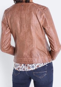 Person med en åtsittande brun läderjacka över en blommig skjorta och mörkblå jeans, vänd bort från kameran mot en enkel bakgrund.