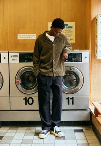 Jeune homme vêtu de vêtements décontractés se tient devant des machines à laver numérotées 10 et 11 dans une laverie à panneaux de bois.