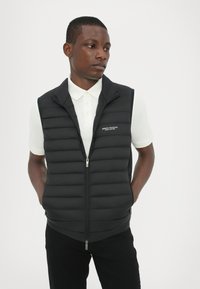 Sort polstret vest med lynlås foran, høj krave og to sidelommer. Har et logo på venstre bryst og en struktureret quiltet design.