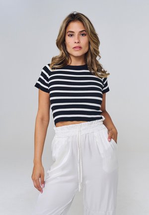 Woman with wavy hair wearing a black and white striped crop top and white high-waisted loose pants against a plain background.