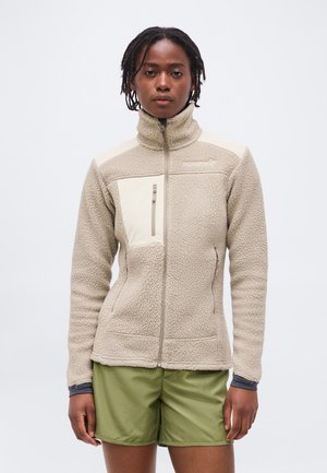 Γυναίκα με κοντά dreadlocks που φοράει μπεζ φλις μπουφάν με τσέπη στο στήθος με φερμουάρ και χακί σορτς, στέκεται μπροστά σε λευκό φόντο.