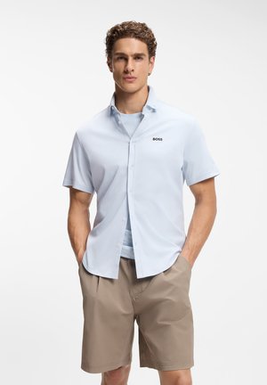 Mężczyzna w jasno-niebieskiej koszuli z krótkim rękawem z logo "BOSS" oraz beżowych szortach, stoi z rękami w kieszeniach na białym tle.