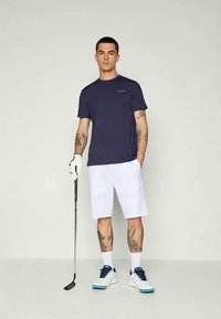 Tengerészkék sport póló, fehér rövidnadrág és fehér golfcipő tengerészkék részletekkel; golfütőt tart; karjain tetoválásokkal.