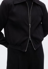 Μαύρο μπουφάν μπόμερ με φερμουάρ στο μπροστινό μέρος, διαθέτει ριμπ μανσέτες και τελείωμα. Κατασκευασμένο από απαλό ύφασμα, έχει δομημένο γιακά και μίνιμαλ σχεδιασμό.