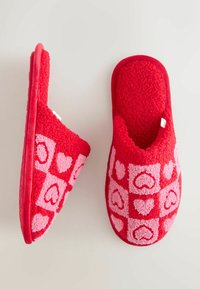 Červené chlpaté papuče s ružovými vzormi srdiečok, otvoreným zadkom, jemnou textúrou, polstrovanou podrážkou a zakriveným dizajnom pre pohodlie.