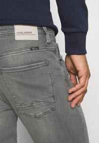 Šedé džínové kalhoty s texturovaným povrchem, s koženou nášivkou označenou "BLEND" a detailem boční kapsy.