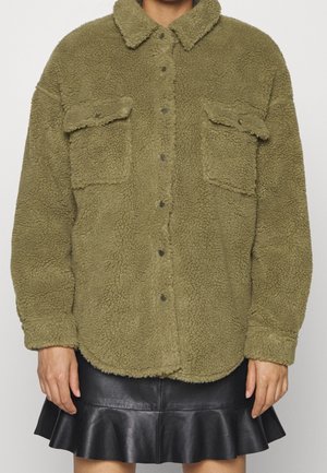 Olivengrøn sherpa fleecejakke med knaplukning og to brystlommer med klap, båret over et sort lædernederdel med flæser.