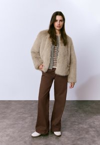 Mujer joven con cabello largo castaño, vestida con una chaqueta de piel sintética beige, una camiseta de rayas, pantalones de pierna ancha marrones y zapatillas blancas, de pie sobre un suelo gris.