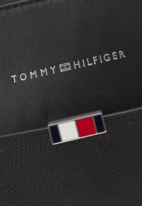 Sort læderpung med tekstureret overflade, der har et sølvfarvet "Tommy Hilfiger" logo og en rektangulær multifarvet detalje.