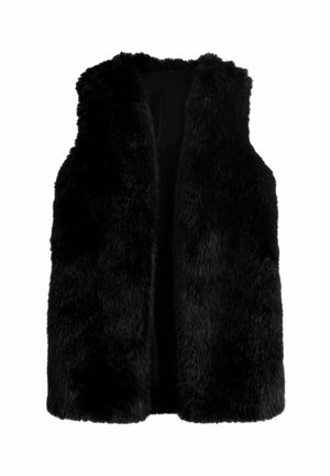 Person trägt eine schwarze Kunstfellweste über einer schwarzen Lederjacke mit blauen Jeans, steht seitlich an einer hellen Wand.