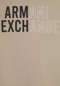 T-shirt in cotone beige con grande scritta nera e bianca "ARMANI EXCHANGE", con motivi a griglia in una tonalità più chiara sullo sfondo.