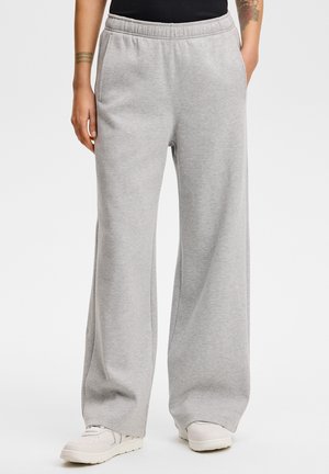 Persoon die lichtgrijze wijde joggingbroek met elastische taille en witte sneakers draagt, staand tegen een effen lichte achtergrond.