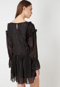 Vestido negro con una tela texturizada y brillante. Presenta acentos de volantes en los hombros y las mangas, y una abertura tipo ojal en la parte posterior.