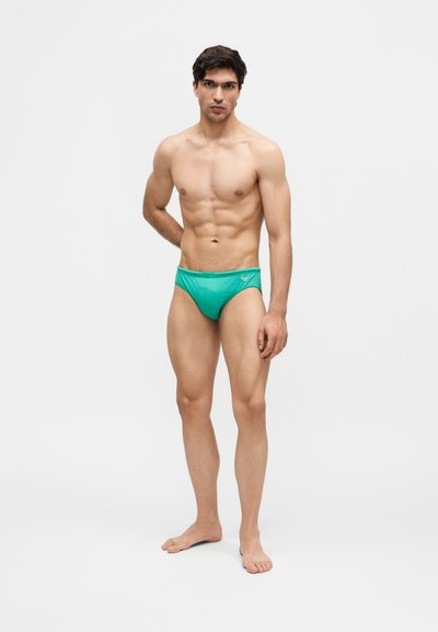 Νέος μυώδης άνδρας που φοράει τιρκουάζ μαγιό, στέκεται ξυπόλητος μπροστά σε απλό φωτεινό φόντο με το ένα χέρι πίσω από την πλάτη.