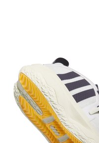 Vit sportskor med marinblå ränder, texturerad gummisul med gula och vita detaljer, framträdande häldesign och varumärkesdetaljer på sidan.