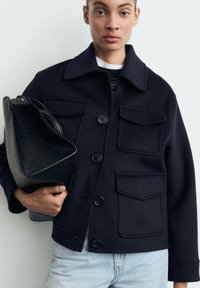 Donna che indossa un giacchetto di lana blu scuro con grandi bottoni e tasche a pattina, mentre tiene una borsa in pelle nera, abbinata a jeans azzurri chiari.