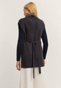 Charcoal gray knit vest with open front, ribbed hem, and waist tie. Features a textured pattern and wide armholes, layered over a black top.