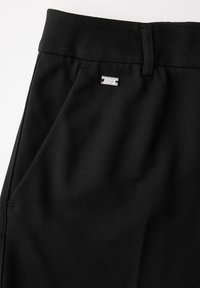 Pantalon noir à texture lisse, avec une poche avant, une étiquette de marque discrète et une ceinture repliée.