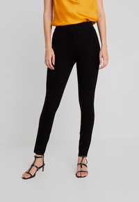 Černé Legíny s vysokým pasem, s přiléhavým střihem a vertikálními švy, doplněné černými sandály na podpatku a žlutým topem.